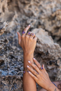 Model wearing five Violet Horizon Amethyst crystal rings in gold, set against a rocky backdrop, showcasing the raw beauty of the crystals.