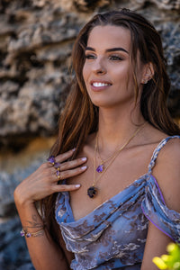 Model adorned with a Violet Horizon Amethyst crystal necklace featuring large and small amethyst stones set in gold, complemented by matching amethyst rings and bangles, all beautifully framed against a soft, blurred rock backdrop.