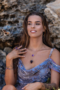 The model, set against a blurred rocky backdrop, wears a Violet Horizon Amethyst crystal necklace with large and small amethyst stones in gold, complemented by amethyst rings and bangles.