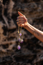 Close-up of model holding four Violet Horizon Amethyst Crystal Necklaces in gold, with a slightly different angle, highlighting the raw, hand-dug amethyst stones against a blurred rock background.