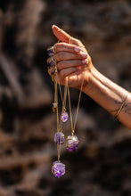 Close-up of a model holding four Violet Horizon Amethyst Crystal Necklaces in gold, with a blurred rocky background.