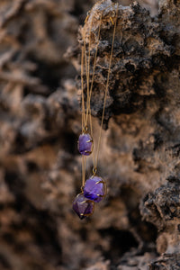 Three Violet Horizon Amethyst crystal necklaces in gold, featuring amethyst stones, draped over a rock surface, creating a textured, earthy display.