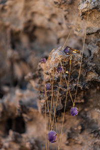 Violet Horizon Amethyst crystal necklaces in gold, draped over rocks with the Blackwood Ave logo visible. Amethyst rings are also displayed, adding a touch of elegance to the natural setting.