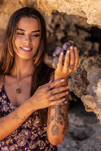 Close-up of the model holding a raw amethyst cluster in her hands, set against a rocky backdrop, while wearing the Twilight Veil Amethyst Crystal Cluster Necklace in gold around her neck.